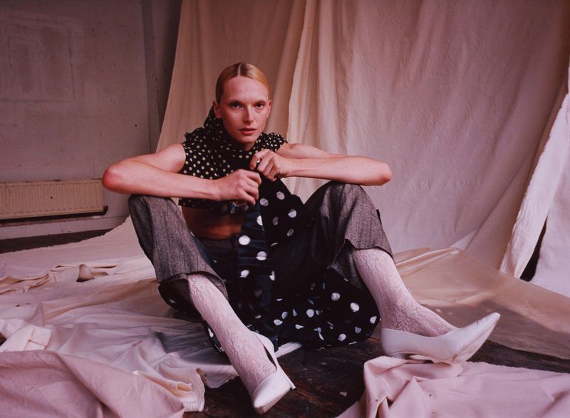 A model poses on a white sheet, wearing grey trousers, white heels and socks, and a polka dot top and headscarf in a studio.