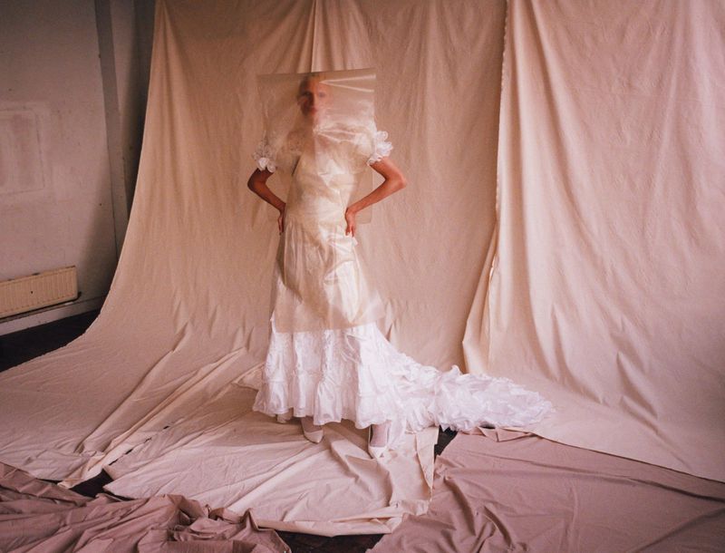 Woman in white gown with fabric over face poses in front of draped fabric backdrop in a studio setting, creating a surreal fashion image.