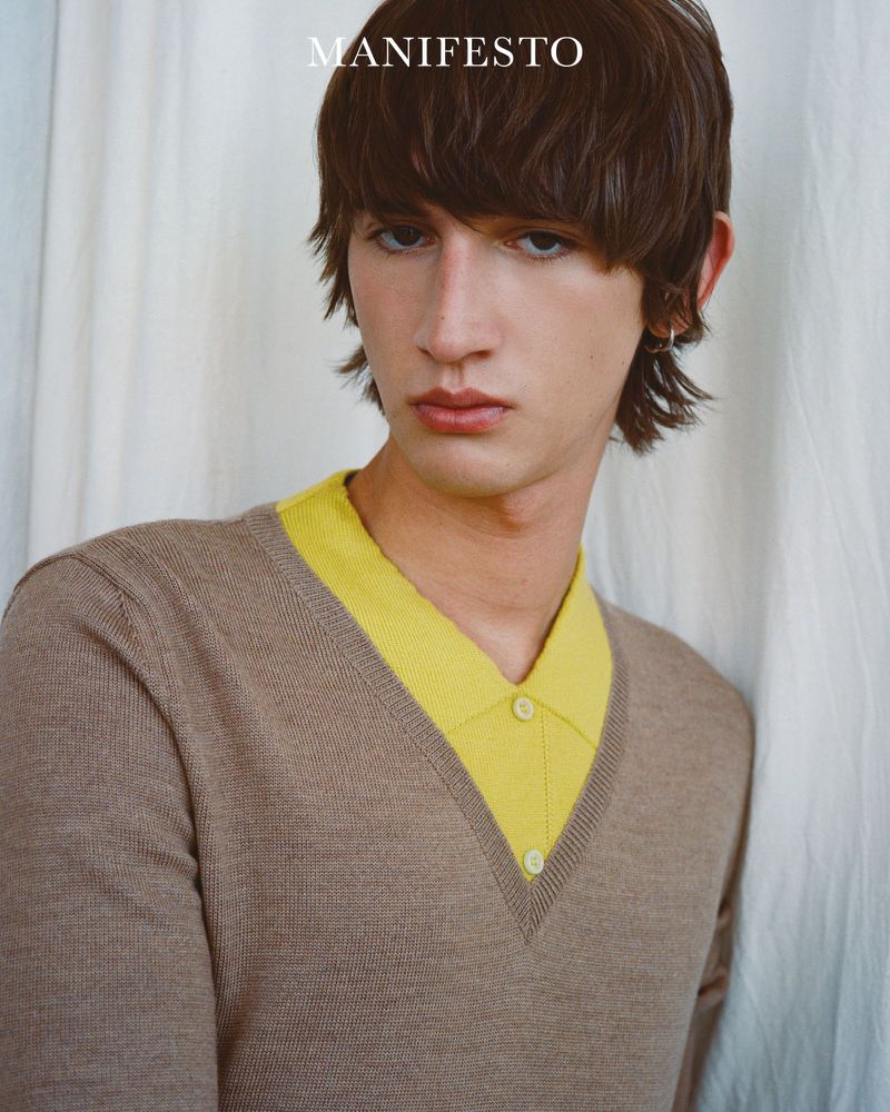 Close-up studio portrait of a young adult with brown hair wearing a beige V-neck sweater with yellow collared shirt underneath, looking at camera.