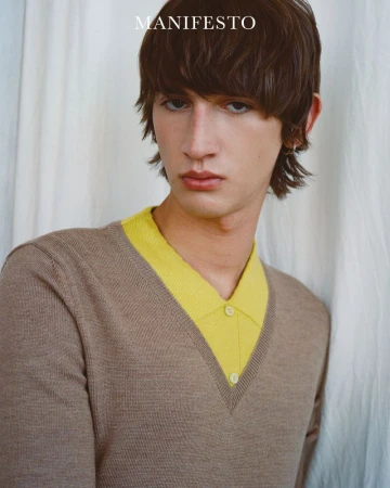 Close-up studio portrait of a young adult with brown hair wearing a beige V-neck sweater with yellow collared shirt underneath, looking at camera.