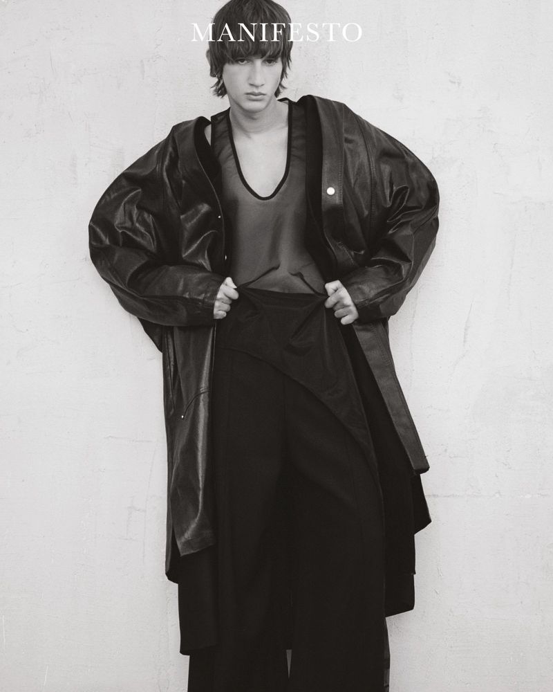A young adult poses in a black mesh top, wide-legged pants, and an oversized leather jacket against a textured white wall in black and white.