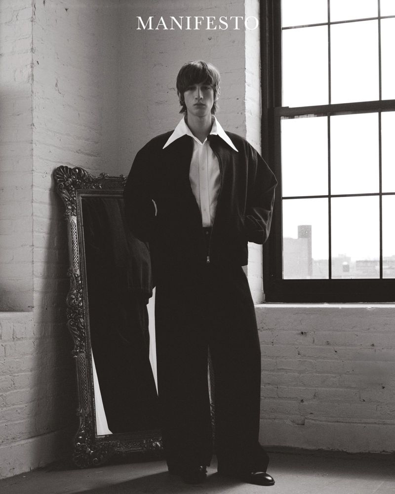 A young adult man in a dark jacket and wide-leg trousers stands near a window and ornate mirror in a minimalist indoor studio setting.