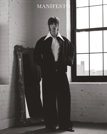 A young adult man in a dark jacket and wide-leg trousers stands near a window and ornate mirror in a minimalist indoor studio setting.