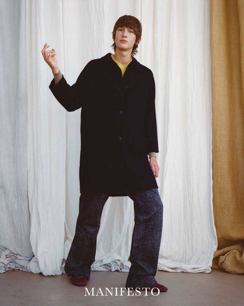 A long-haired male fashion model in a black coat and wide-leg patterned trousers poses in front of a neutral-toned draped fabric backdrop.