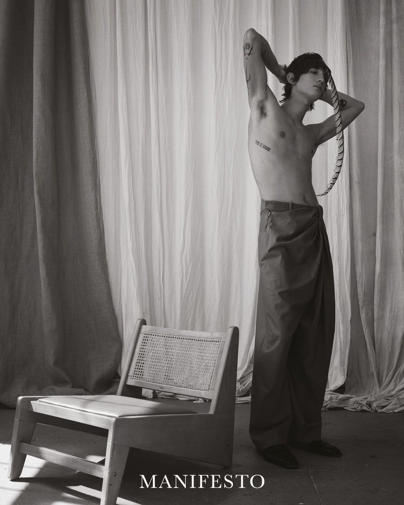 Monochrome studio shot of shirtless male model with tattoos posing next to a vintage chair and draped fabric background, high-fashion editorial.