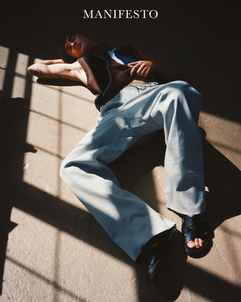 A young adult lying on the floor in a sunlit interior, wearing a brown top, light blue pants, and sandals, with 'MANIFESTO' text overlay.