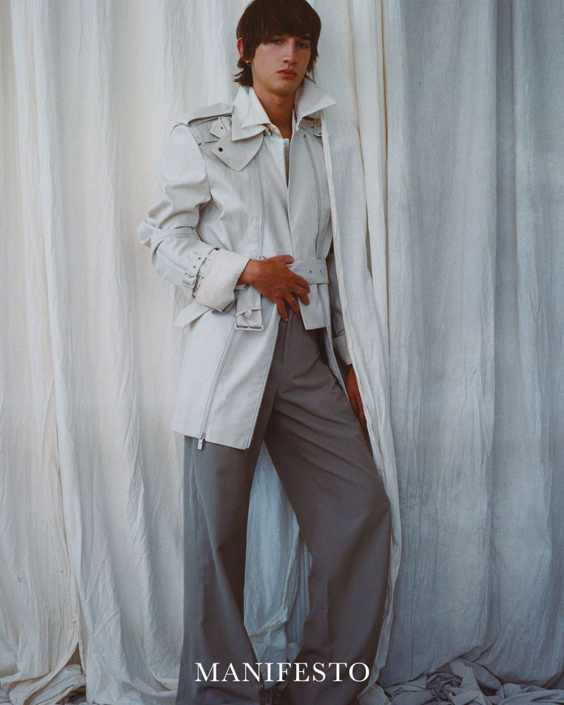 A young adult man poses in an open white jacket and grey wide leg trousers against a draped white fabric backdrop in a studio shot.