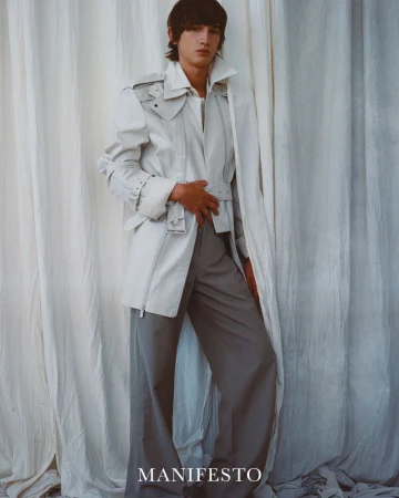 A young adult man poses in an open white jacket and grey wide leg trousers against a draped white fabric backdrop in a studio shot.
