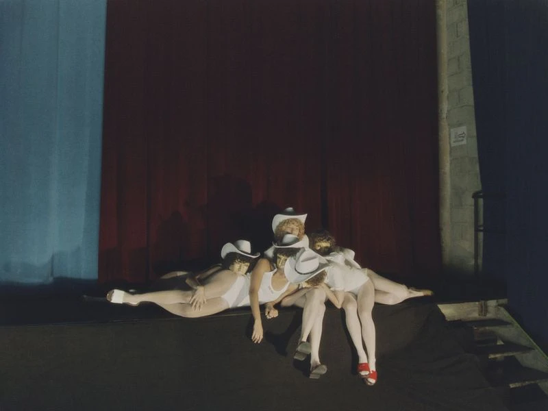 Three performers in white bodysuits and cowboy hats pose on a dark stage with a red curtain backdrop and minimal lighting.