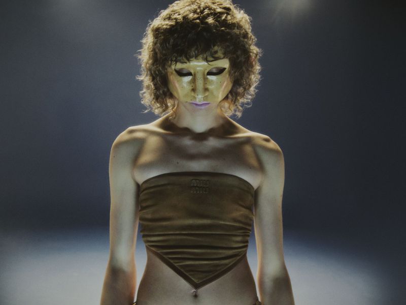 Adult female with a tan tube top and gold mask stands in a spotlight, shot at eye level, emphasizing the texture and form of her clothing.