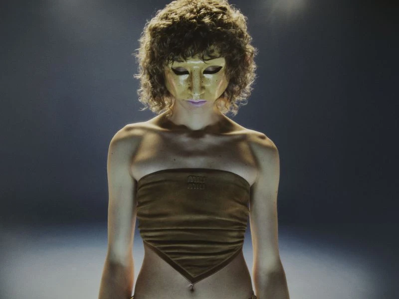 Adult female with a tan tube top and gold mask stands in a spotlight, shot at eye level, emphasizing the texture and form of her clothing.