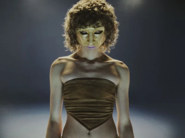 Adult female with a tan tube top and gold mask stands in a spotlight, shot at eye level, emphasizing the texture and form of her clothing.
