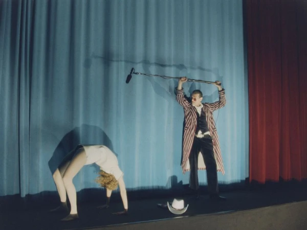 Performer in bridge pose and stagehand with boom mic in front of a blue and red curtain create a vintage theatrical scene.