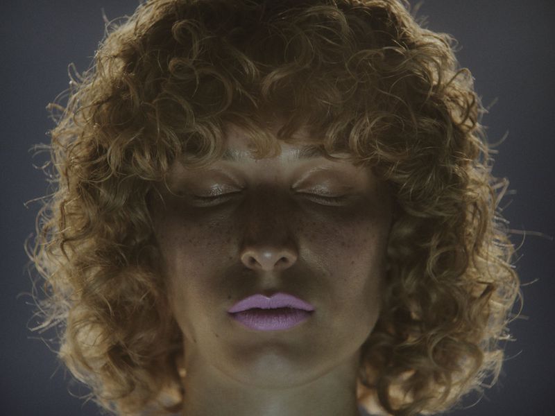 Portrait of a woman with curly hair and closed eyes in soft lighting, showcasing beauty and serenity against a dark, smooth background.