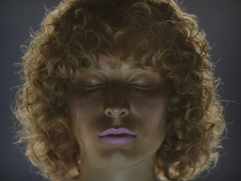 Portrait of a woman with curly hair and closed eyes in soft lighting, showcasing beauty and serenity against a dark, smooth background.