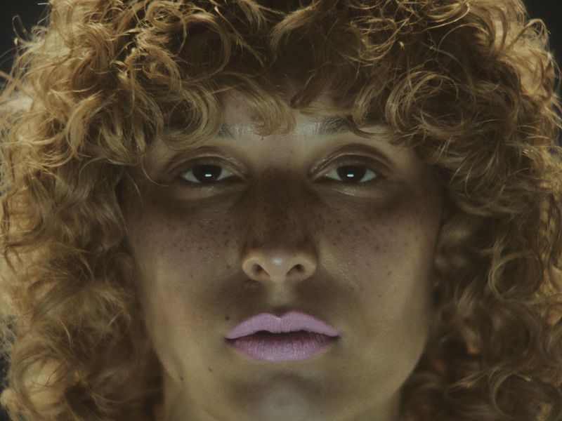 Close-up portrait of a model with textured curly hair, freckles across the nose, and pale lilac lipstick against a dark background.