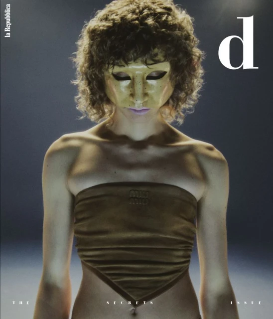 Magazine cover featuring a person with curly hair wearing a gold mask and a brown Miu Miu top, against a blurred, dark background.