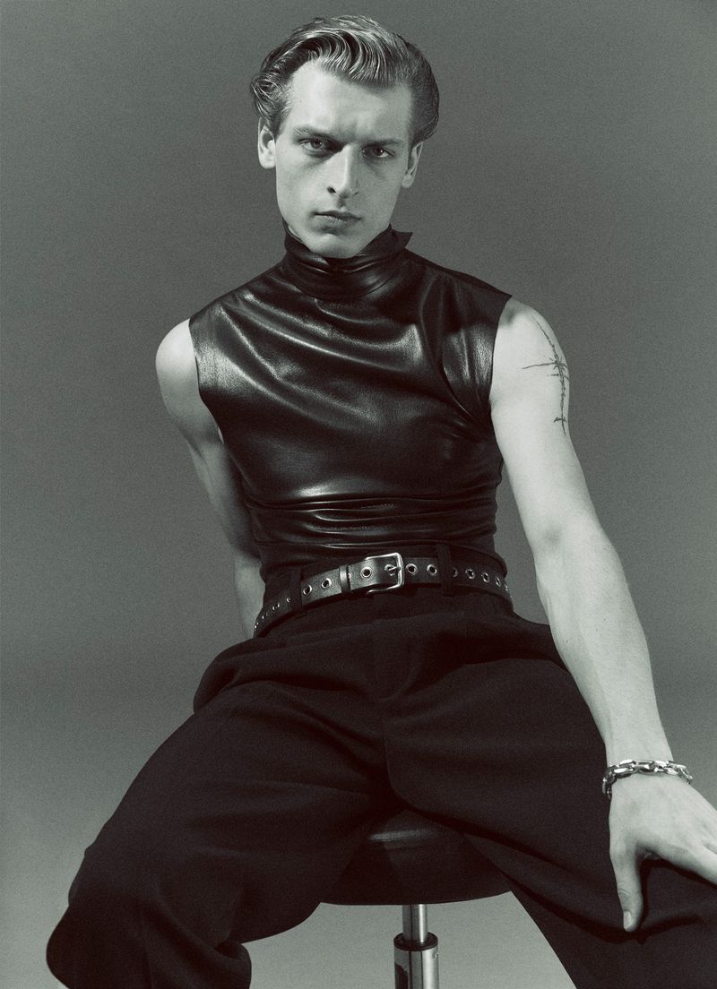 Monochrome studio portrait of a fair-skinned adult male wearing a black leather top and pants while seated on a stool.
