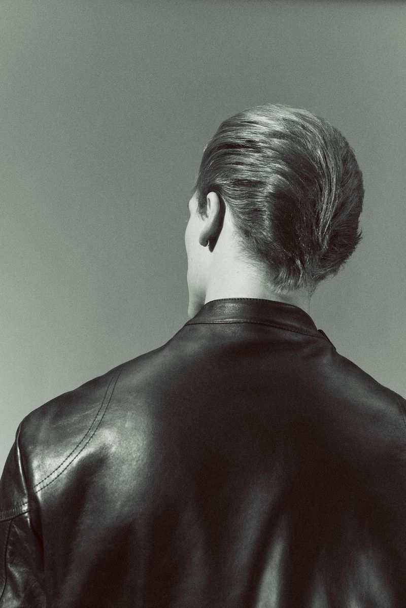 Back of an adult male with blond hair wearing a black leather jacket against a gray background in a portrait-style, eye-level shot.
