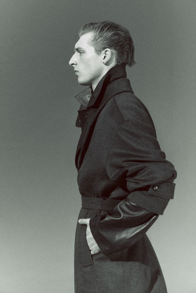 Monochromatic portrait of a man in a dark wool coat with leather sleeves, looking to the left, in a photo studio against a gray background.