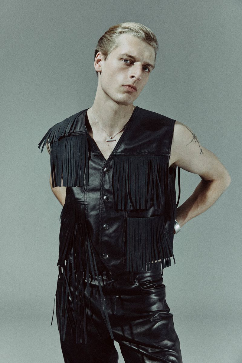 Stylish blond male model poses in a black leather fringe vest and matching pants against a grey background in a studio shot.