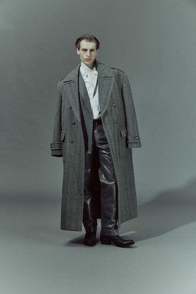A full-length studio shot showcases a male model in a grey herringbone overcoat, vest, dress shirt, black leather trousers and boots.