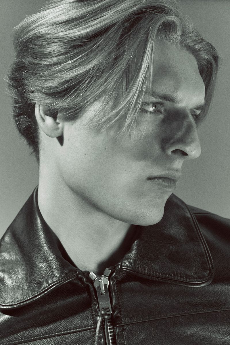 Close-up profile of a man with styled blonde hair wearing a black zipped leather jacket against a neutral studio backdrop.