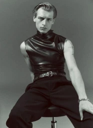 Model Colin Otto seated wearing a sleeveless black leather turtleneck and grommet belt against a grey studio backdrop.
