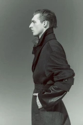 Profile portrait of a male model in a dark belted coat against a neutral studio background for Wallpaper Magazine