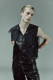 Model wearing a black fringed leather vest and leather trousers against a grey studio backdrop