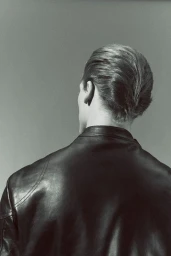 Rear view portrait of a man with styled slicked-back hair wearing a black leather jacket against a seamless grey backdrop.