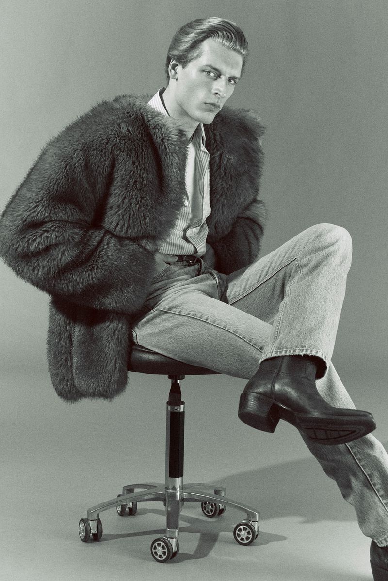 Monochrome fashion editorial portrait of a male model seated on a rolling stool wearing a furry jacket and boots