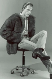 Monochrome fashion editorial portrait of a male model seated on a rolling stool wearing a furry jacket and boots