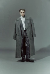 Model Colin Otto wearing a long grey overcoat and black leather pants against a solid grey studio backdrop.