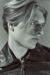 Close-up profile of a man with styled blonde hair wearing a black zipped leather jacket against a neutral studio backdrop.