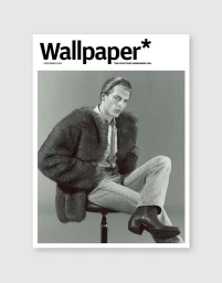 Monochrome fashion editorial portrait of a male model seated on a stool wearing a textured fur coat and leather boots against a grey studio backdrop.
