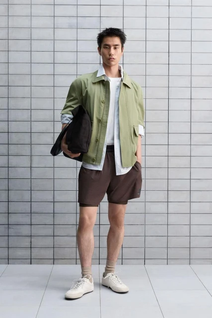 Full shot of an adult Asian male model in a sage-green jacket, shorts, sneakers, holding a black bag, posing in a photo studio against a tiled wall.