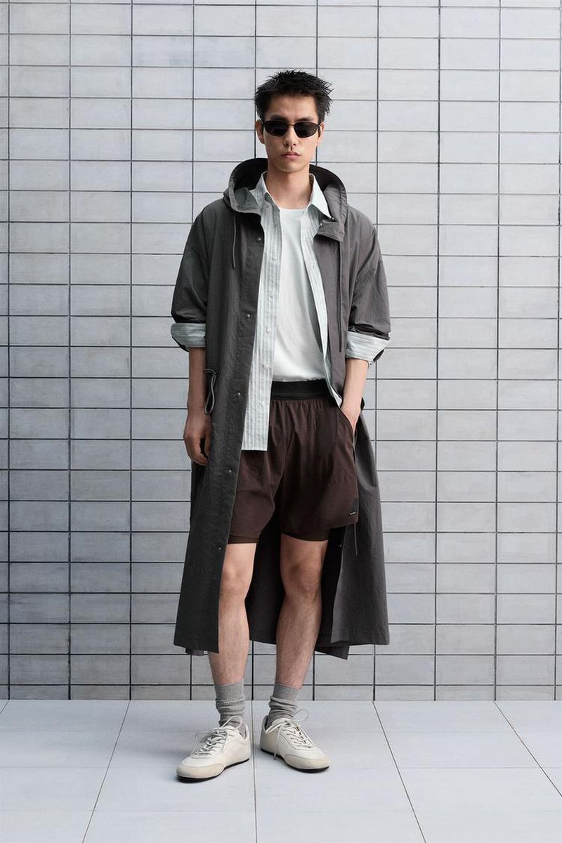 A stylish Asian man posing in a gray hooded coat and brown shorts, complemented by white sneakers against a tiled wall backdrop.