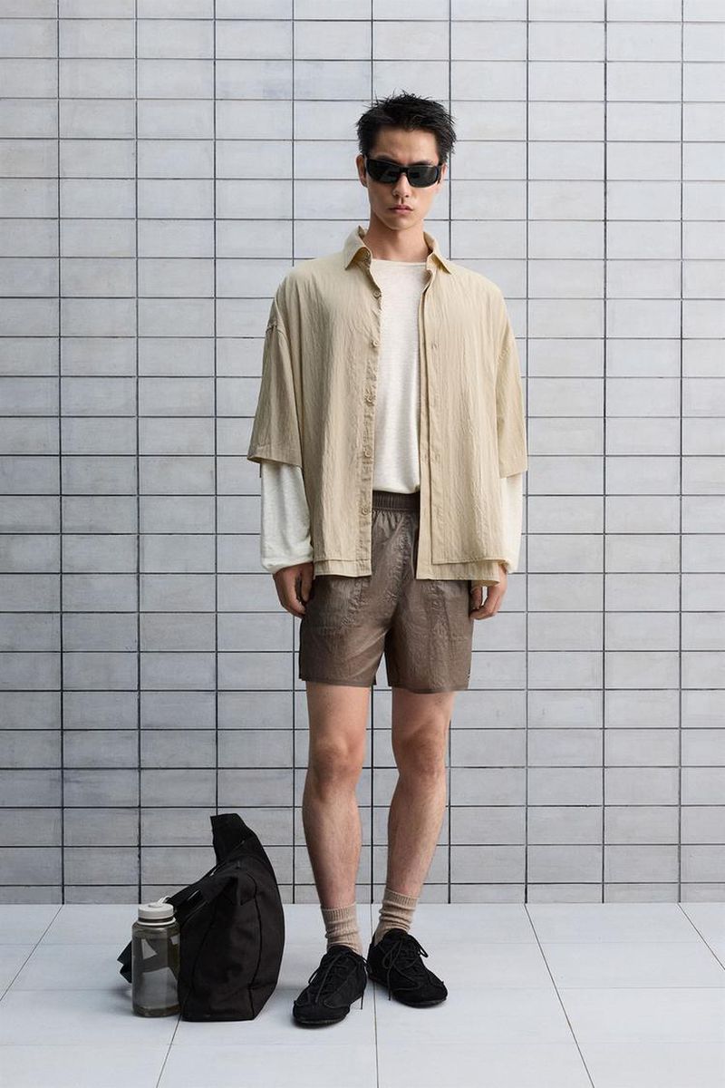 A young adult man stands in a photo studio wearing a beige button-down shirt, a white t-shirt, brown shorts, sunglasses, and black sneakers.