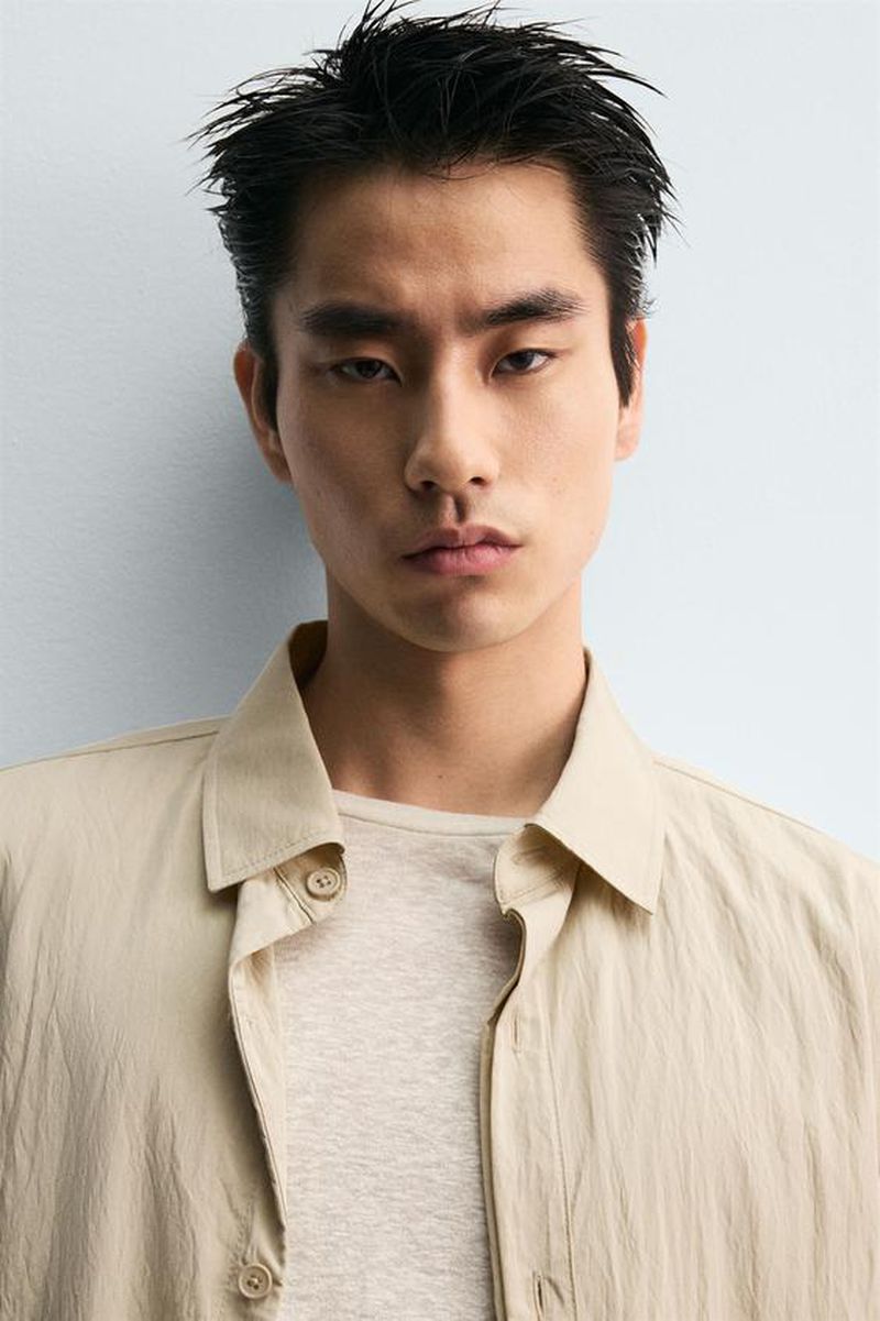 Portrait of a young adult male with dark hair, wearing a beige button-up shirt and t-shirt, against a neutral background.
