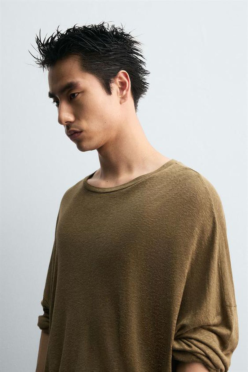 Headshot of a young Asian man with spiky black hair wearing a loose, tan-colored shirt against a neutral background in a studio setting.
