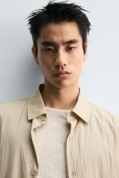 Close-up portrait of a male model wearing a beige button-down shirt against a light grey background.