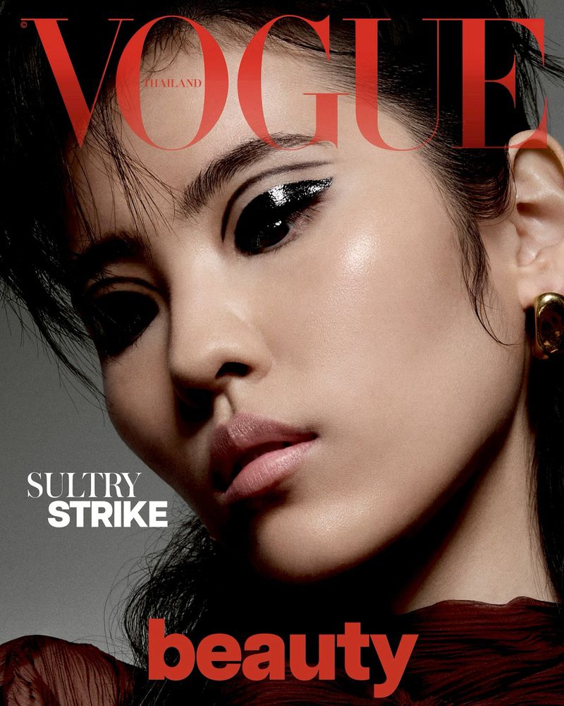 Close-up portrait of a woman with dramatic black eyeliner, glossy skin, and gold earrings. Vogue Thailand logo visible at the top.