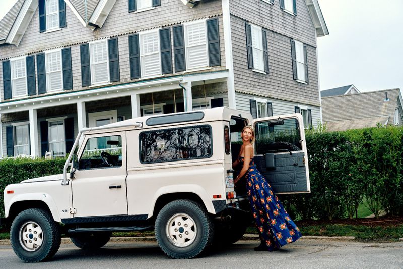 Blonde woman in a floral dress leans against an open door of a Land Rover SUV near a grey house with black shutters.