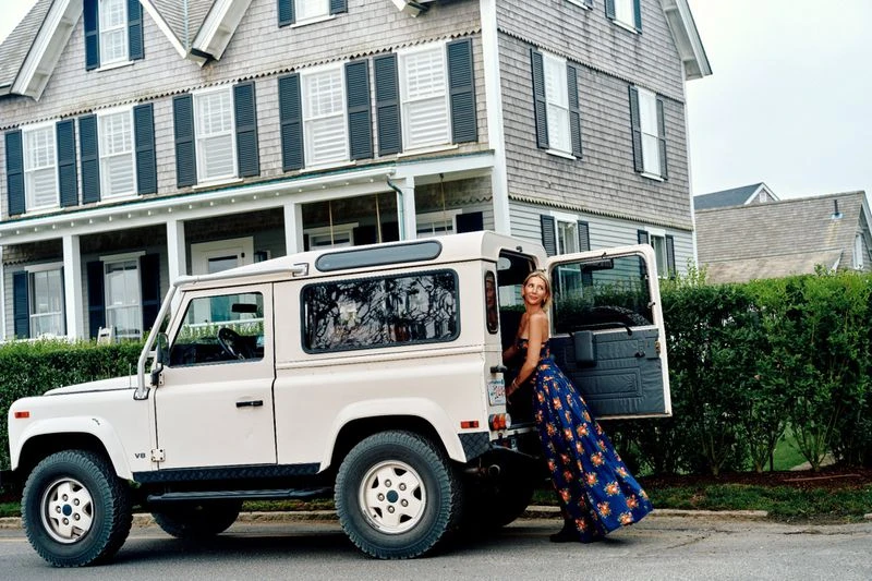 Blonde woman in a floral dress leans against an open door of a Land Rover SUV near a grey house with black shutters.