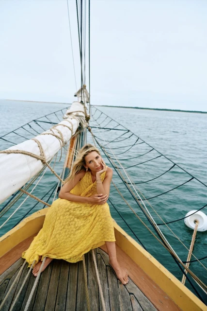 Woman in a yellow lace dress poses on the bow of a sailboat on the water on a cloudy day, fashion shoot, travel photography.