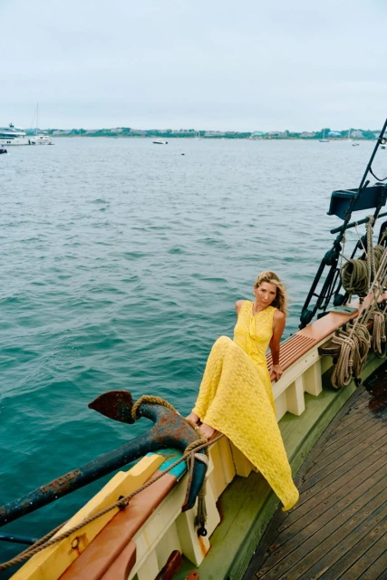 A woman in a yellow dress poses on a boat, leaning against an anchor with the sea and a distant shoreline in the background on a cloudy day.