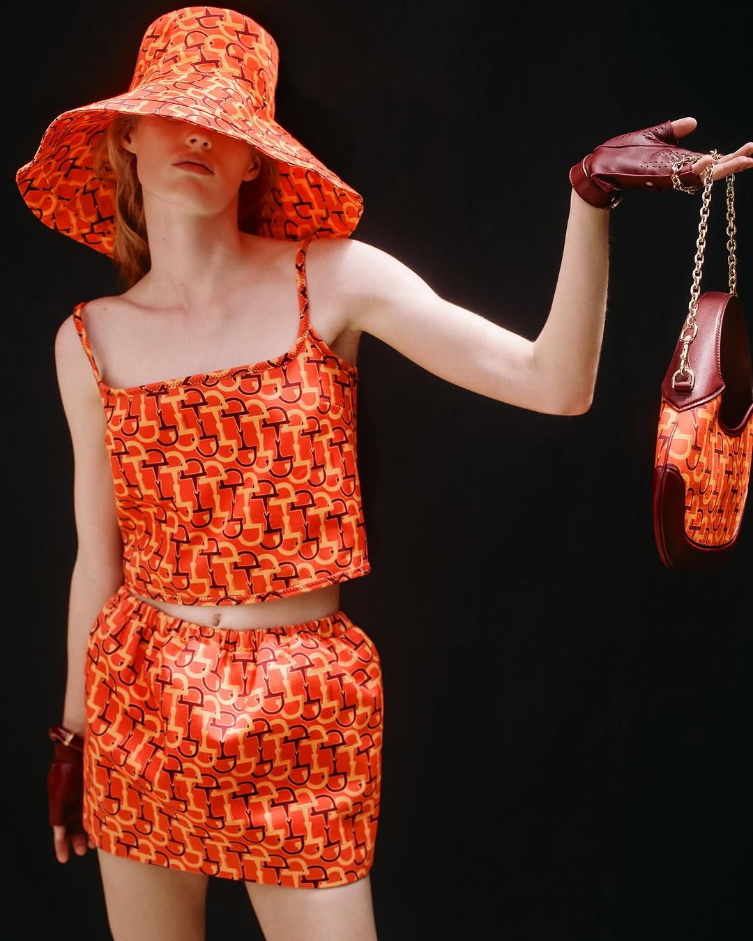 Woman poses in a studio, wearing a printed orange two-piece set with matching hat and handbag against a dark background.