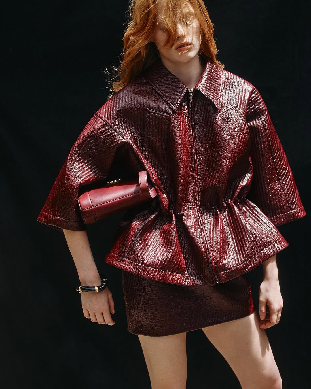 Woman in a shiny burgundy jacket and matching skirt. A matching burgundy purse is positioned at her waist against a dark background.