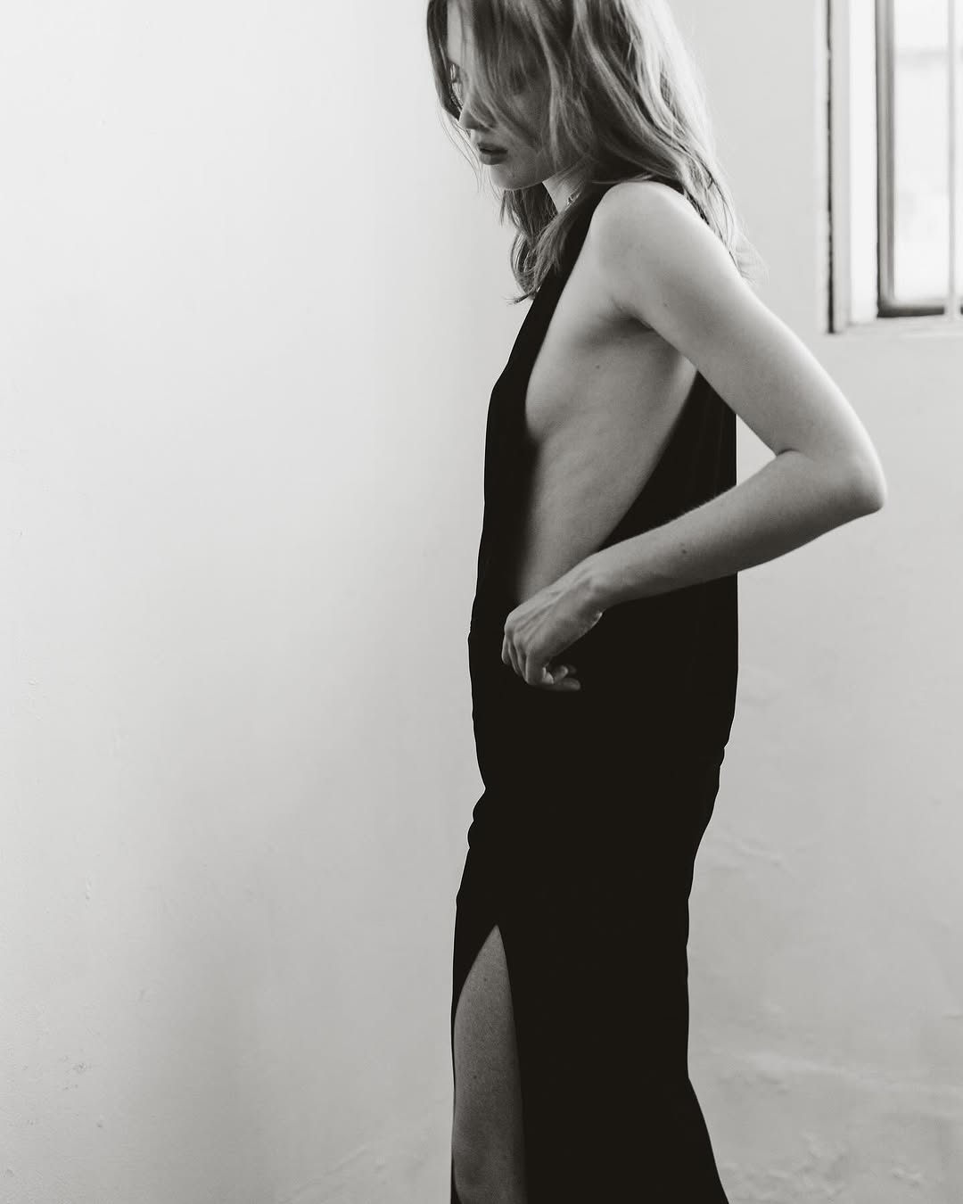 A slender woman with blonde hair poses in profile wearing a black, sleeveless dress with a thigh-high slit against a white wall.